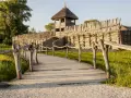Drewniana kładka prowadzi do grodziska, jednego z historycznych miejsc w Polsce. Widać palisadę i wieżę obronną.