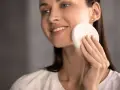 Une femme souriante utilise une éponge naturelle pour se laver le visage. Une méthode douce pour un nettoyage en douceur.