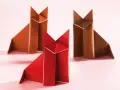 Drei origami Füchse, einer rot, zwei braun, sitzen auf rosa Untergrund.