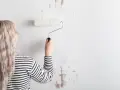 Mujer con cabello rubio y camiseta a rayas está a punto de empapelar pared con rodillo blanco.