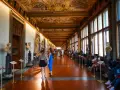 Wnętrze muzeum Uffizi z rzeźbami i malowanym sufitem. Ludzie spacerują i odpoczywają wzdłuż korytarza.