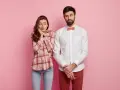 Coppia su sfondo rosa. Lei, con camicia a quadri e jeans, sembra timida. Lui, con camicia bianca e papillon, è pensieroso. Come vestirsi al primo appuntamento uomo?