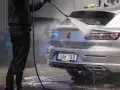 En person tvättar en silverfärgad bil med högtryckstvätt. Frågan "får man tvätta bilen hemma" dyker upp när man ser detta.