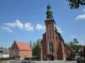 Majestatyczna kolegiata w Kartuzach z cegły, z zieloną wieżą i zegarem, na tle błękitnego nieba z chmurami.