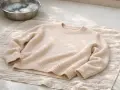 Un pull en laine beige est posé sur une serviette, prêt à laver la laine. Un bol d'eau savonneuse est à proximité.