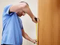 Mężczyzna w niebieskiej koszulce mierzy drzwi taśmą mierniczą.