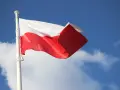 Biało-czerwona bandera polska powiewa na maszcie na tle błękitnego nieba z chmurami.