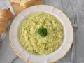 Kremowe risotto z brokułami, posypane pistacjami, z małą różyczką brokułu na wierzchu. Obok widelca i kawałków parmezanu.