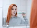 Rudowłosa kobieta myje zęby szczoteczką soniczną, zastanawiając się, czy szczoteczka soniczna usuwa kamień.