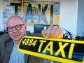 Mann mit Brille hält ein gelbes Taxi-Schild mit der Nummer 4884. Im Hintergrund Kunstwerke mit "TAXI" und der Zahl 1884.