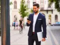 Mężczyzna w eleganckim stroju biznesowym męskim sprawdza telefon, trzymając laptopa.
