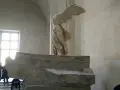 Skrzydlata Nike, symbol zwycięstwa, niczym mit, unosi się nad kamiennym postumentem.