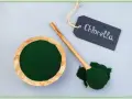 Chlorella w proszku w drewnianej miseczce i na łyżce. Tabliczka z napisem "Chlorella" podpowiada, jak stosować ten zielony proszek.