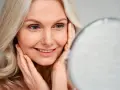 Une femme blonde sourit en se regardant dans un miroir, ses mains caressant son visage. Le maquillage paupière tombante avant après est subtil.