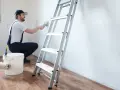 Hombre con overol azul y gorra, usando brocha para limpiar paredes. Escalera y cubo de pintura a su lado.
