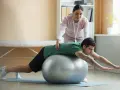 Fizjoterapeutka wspiera mężczyznę w ćwiczeniach na piłce. Rekonwalescencja po nietrzymaniu moczu, przygotowanie do powrotu do pracy fizycznej.