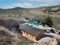 Casa de madera con piscina y caravana en terreno rústico, rodeada de olivos y colinas. Ideal para quienes buscan un refugio natural.