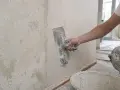 Ręka nakłada gładź polimerową na ścianę za pomocą kielni. Widać starą, łuszczącą się farbę i wiadro z masą.