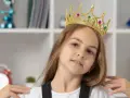Mała egocentryczka w złotej koronie z klejnotami, pewna siebie, z uśmiechem na twarzy, na tle regału z książkami i zabawkami.