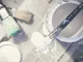 Pędzel z białą farbą winylową na gazecie, obok otwarty pojemnik z farbą i inne pędzle.