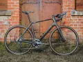A sleek black Salsa Warroad gravel bike with orange accents stands against a rustic brick wall and weathered metal door.