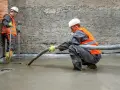 Dwóch budowlańców w kamizelkach odblaskowych wylewa pianobeton. Jeden kieruje wężem strumień materiału, drugi kontroluje poziom.