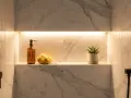 Une niche dans la salle de bain, éclairée par une lumière douce, présente une bouteille de savon, une éponge naturelle et une petite plante verte.