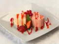 Dwie panna cotta w kształcie serc, polane sosem truskawkowym, ozdobione owocami i miętą.