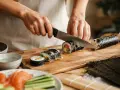 Oto jak się robi sushi: ręce kroją rolkę sushi na desce. Obok ryż, łosoś, awokado i ogórek.