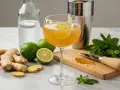 Un verre de boisson dorée garni d'une tranche de citron vert, entouré de gingembre frais, de citrons verts et de menthe. Une préparation pour une infusion gingembre citron rafraîchissante.