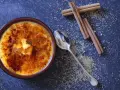 Cremige Crema Catalana mit karamellisierter Zuckerkruste, daneben Zimtstangen und Zucker.