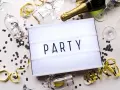 Świętowanie zaczyna się od zaproszeń! Na stole leżą kieliszki, szampan i konfetti, a tablica świetlna głosi "PARTY".