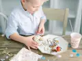 En pojke med en randig skjorta tar en donut med strössel. Det är en festlig dukning med konfetti och en mugg med texten "Hip Hip Hooray". En härlig stund av burken lek.