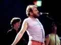 Joe Cocker śpiewa na scenie, a jego charakterystyczne utwory niosą się po sali. W tle widać innych muzyków.