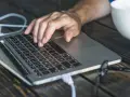 Dłoń na klawiaturze laptopa, przygotowująca się do twardego resetu. Obok biały kubek.