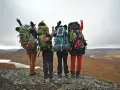 Quatre randonneurs, sacs à dos prêts, contemplent un paysage sauvage. Leur équipement, incluant des panneaux solaires, suggère une préparation pour une liste matériel randonnée 5 jours.