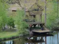 Drewniany domek na palach nad spokojnym jeziorem wiejskim, otoczony zielenią.