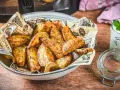 Knusprige Kartoffelecken aus dem Airfryer, serviert in einer Schüssel mit Zeitungspapier. Daneben ein Glas mit Dip und Kräutern.