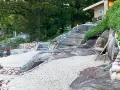 En stenlagd uteplats med grusgångar, stenmurar och trappor som leder upp mot ett hus. En inspiration för en naturlig och robust utomhusmiljö.