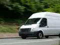 Furgone bianco in movimento su strada, con alberi sfocati sullo sfondo. Potrebbe essere un veicolo soggetto all'articolo 82 del codice della strada.