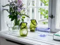 Ein Strauß lila Blumen und grüne Windlichter schmücken die Fensterbank. Ein Glas Wasser und eine blaue Schale runden die Dekoration ab.