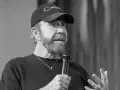 Chuck Norris, mimo swojego wieku, wciąż ma charyzmę. Mówi do mikrofonu w czapce z daszkiem.