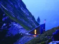 Un moderno bivacco interno, illuminato da una luce calda, si staglia su un pendio montuoso al crepuscolo.