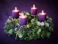 Four purple candles glow on a lush green Advent wreath, adorned with ribbons. The final candle's flame flickers, marking the 4th Advent.