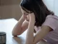 Zmęczona dziewczyna z rękami na skroniach, obok kubek kawy. Paradoksalnie, mimo napoju, czuje senność po kawie.