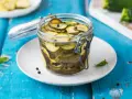 Barattolo di zucchine in agrodolce, con pepe nero e foglie di menta, su un piatto bianco appoggiato su un tavolo blu.