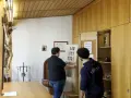 Zwei Männer im katholischen Priesterseminar hängen eine Liste mit Zahlen auf. Ein Kruzifix hängt an der Wand.