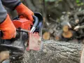 Ręce w pomarańczowych rękawicach trzymają piłę łańcuchową, która przecina pień drzewa. W tle widać stosy drewna, co sugeruje wycinkę drzew w lesie.