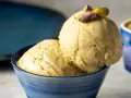 Zwei Kugeln cremiges, gelbes Eis mit Pistazienstückchen, garniert mit ganzen Pistazien, in einer blauen Schale.