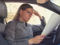 Zmartwiona kobieta w okularach czyta dokumenty w samochodzie, zastanawiając się, co grozi za sprzedaż auta zajętego przez komornika.